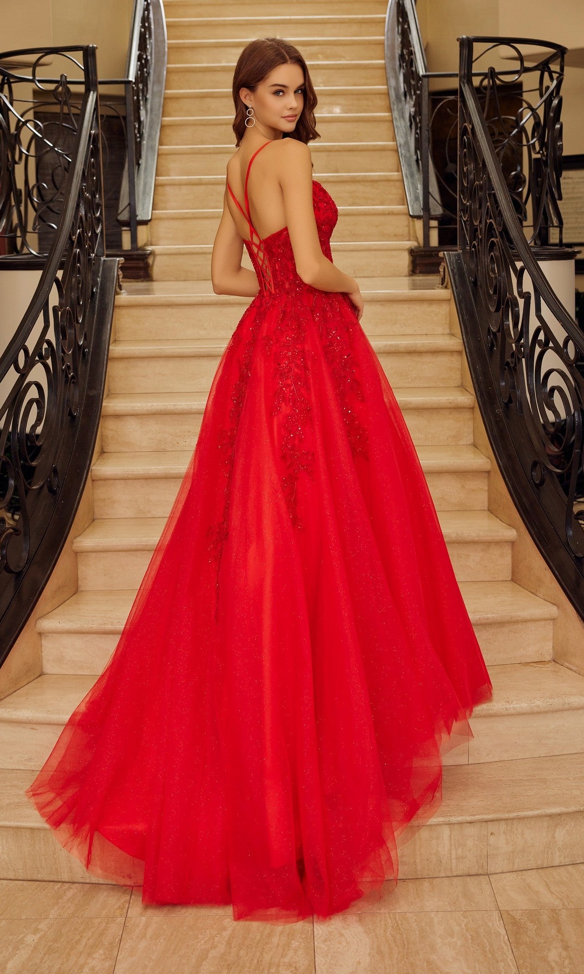 A woman in the Nox Anabel H1883 Long Prom Dress—a red, floor-length gown with a corset bodice and thin straps—stands on a staircase, facing away from the camera.